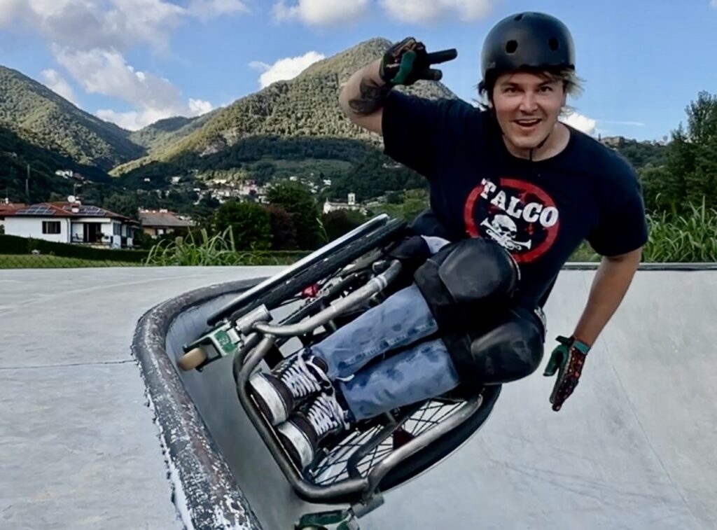 David mit seinem Rollstuhl am carven im oberen Ende einer Corner im Skatepark. Er zeigt das Peace Zeichen, im Hintergrund ein Bergpanorama.
