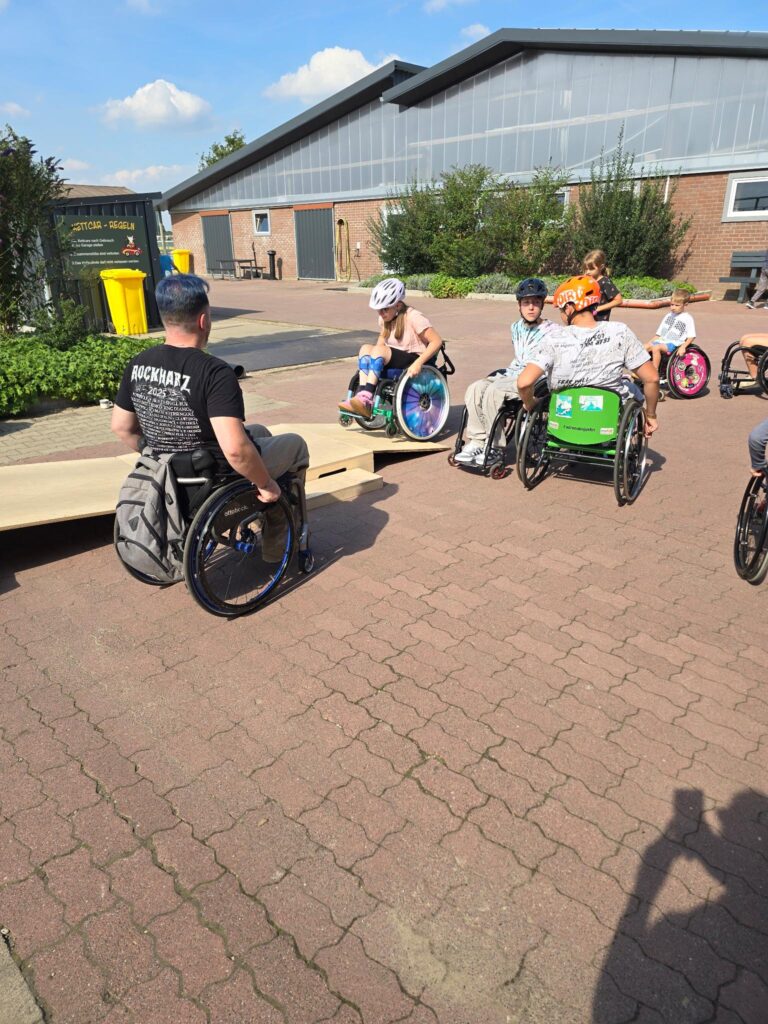 Eine Gruppe von Kindern im Rollstuhl übt das Überwinden einer Rampe auf einem Bauernhofgelände. Ein Trainer im Rollstuhl beobachtet und unterstützt das Geschehen, rundherum herrscht entspannte Atmosphäre.