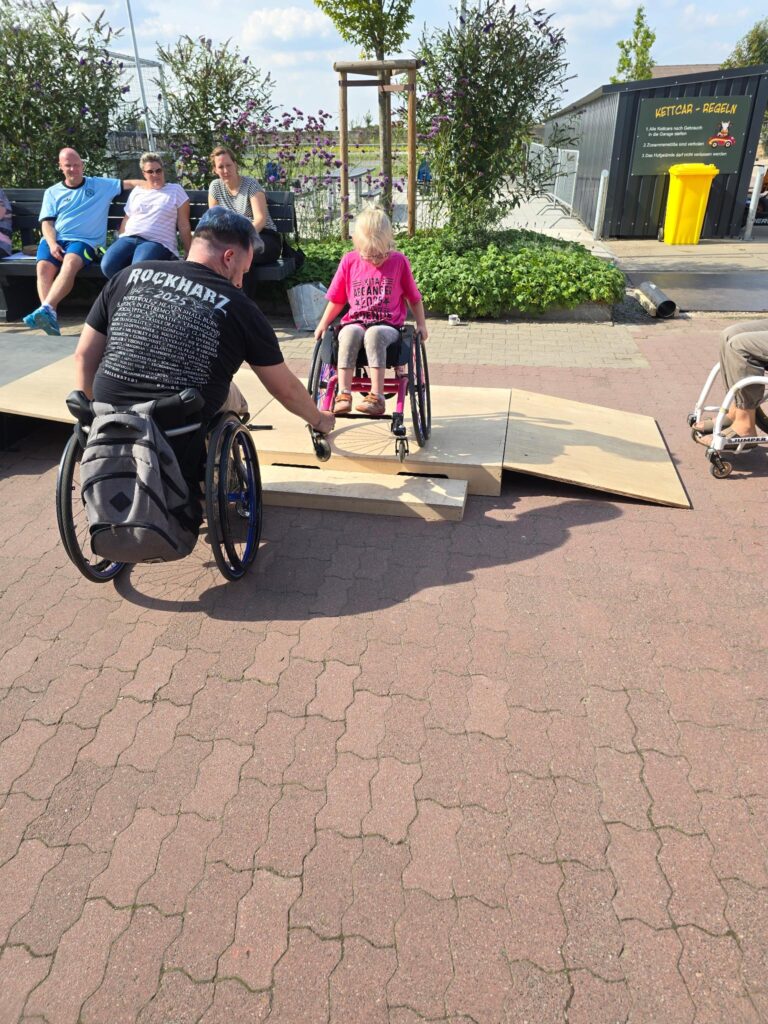 Eine jugendliche Rollstuhlfahrerin mit Helm und bunter Bereifung fährt eine Skate-Rampe hinunter. Ein Mädchen im Rollstuhl und ein Übungsleiter begleiten sie von der Seite. Im Hintergrund sitzen Zuschauer*innen.