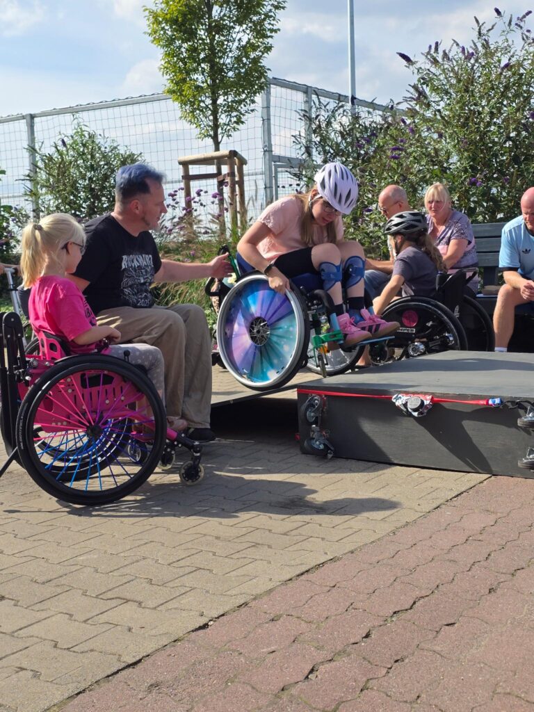 Eine jugendliche Rollstuhlfahrerin mit Helm und bunter Bereifung fährt eine Skate-Rampe hinunter. Ein Mädchen im Rollstuhl und ein Übungsleiter begleiten sie von der Seite. Im Hintergrund sitzen Zuschauer*innen.