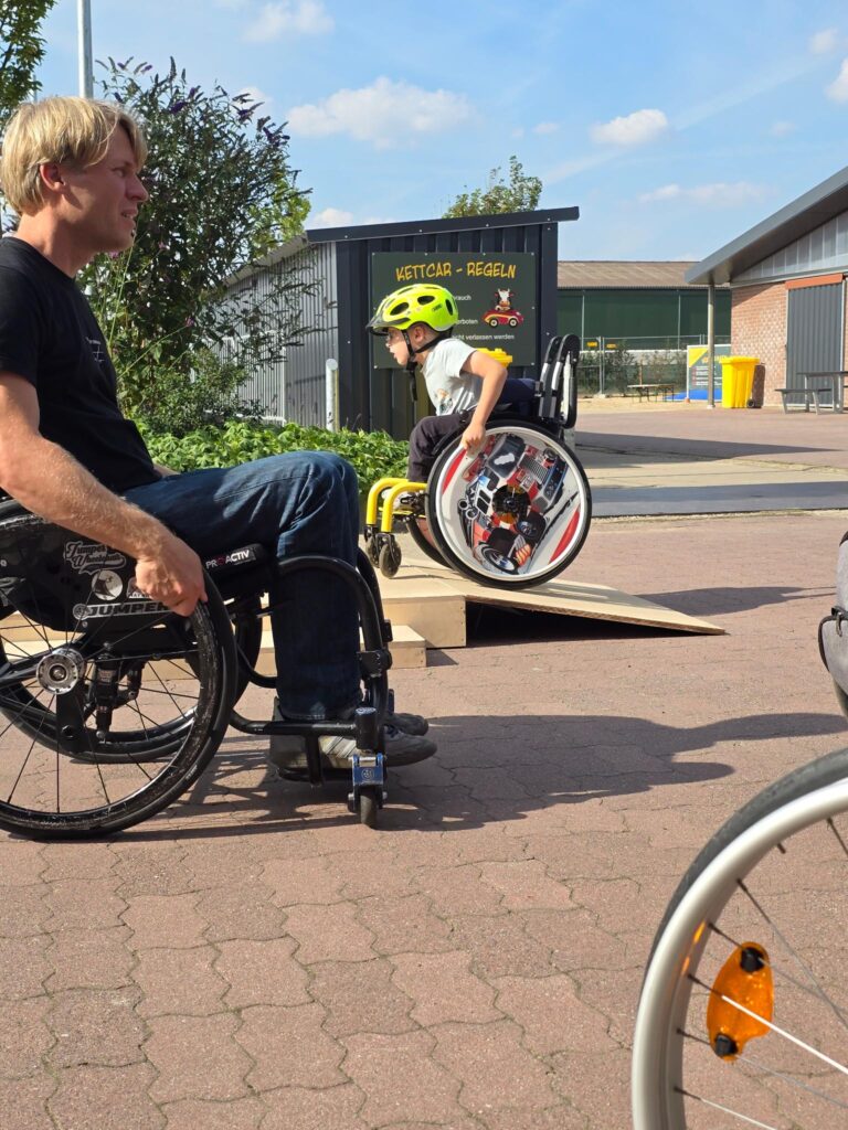 Ein kleiner Junge mit neonfarbenem Helm sitzt im Rollstuhl am oberen Ende einer Rampe und bereitet sich auf die Abfahrt vor. Daneben sitzt ein Erwachsener im Rollstuhl und beobachtet aufmerksam.