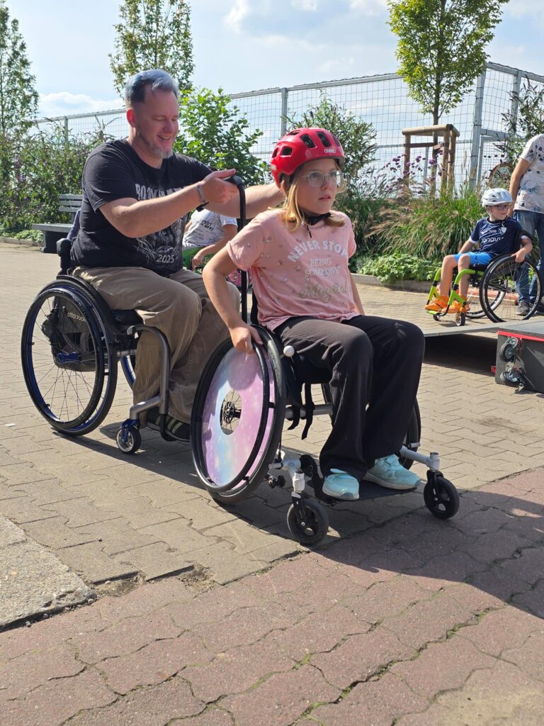 Eine Rollstuhlfahrerin mit blau-violett schimmernden Speichenschützern fährt eine Rampe hinunter. Sie wird von einem erfahrenen Coach angeleitet, daneben sitzt ein junges Mädchen im pinken Rollstuhl.