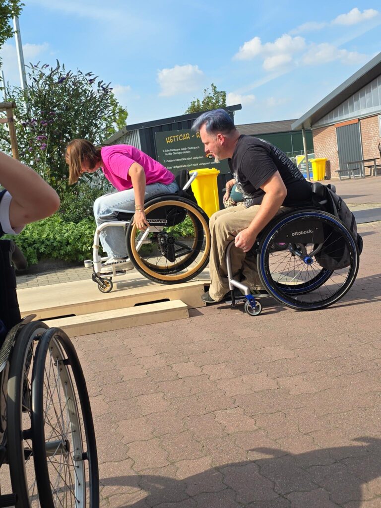 Eine erwachsene Rollstuhlfahrerin fährt auf eine niedrige Plattform, während ein Coach im Rollstuhl neben ihr aufmerksam den Bewegungsablauf beobachtet. Im Hintergrund sitzen weitere Personen auf einer Bank.
