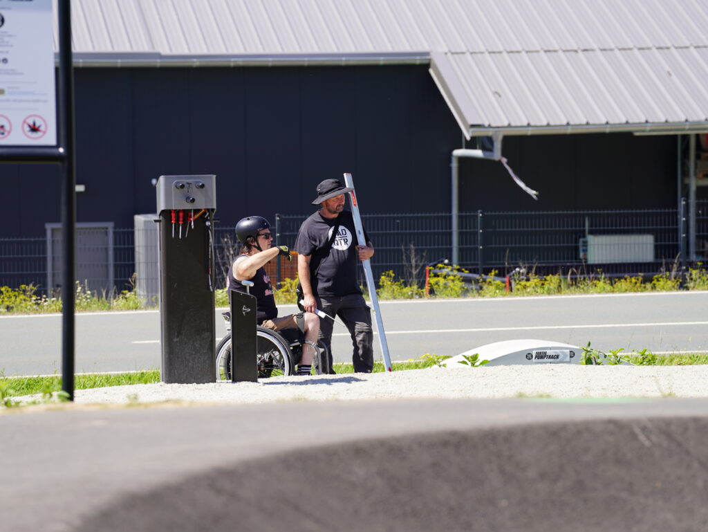 Zwei Männer stehen neben einer Pumptrack und Bikeanlage, einer davon im Rollstuhl. Der Mann im Rollstuhl gestikuliert oder zeigt auf etwas. Der andere Mann hört zu und schaut in die Richtung, die der Rollstuhlfahrer zeigt. Er hält außerdem ein Messinstrument aus Metall.