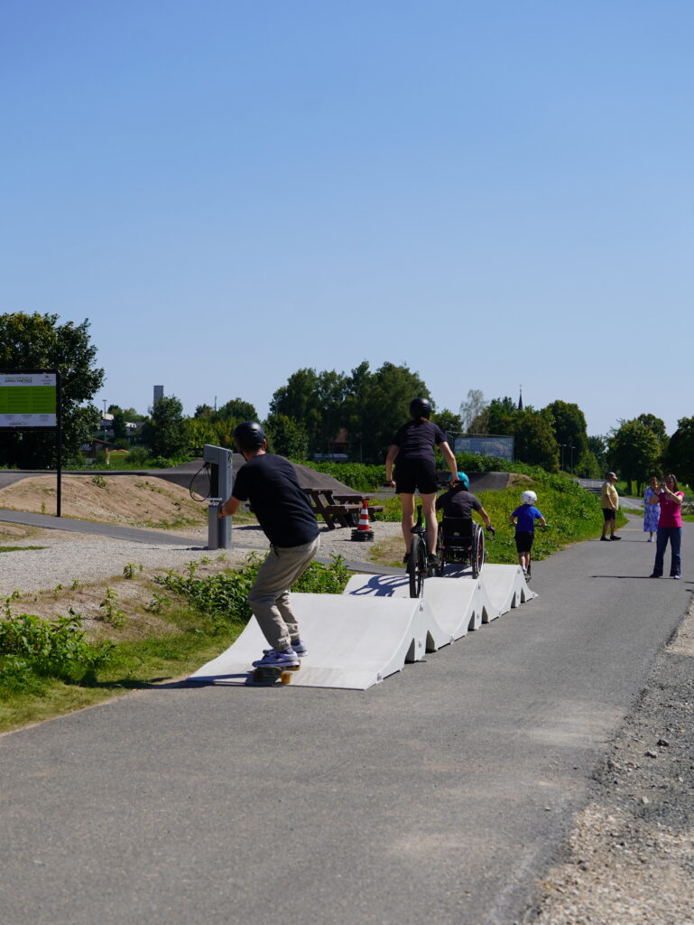 Mehrere Menschen fahren über Kunstoffwellen, die als mobiler Pumptrack auf einen Weg gestellt wurden. Einer fährt Skateboard, ein weiterer Mountainbike, jemand im Rollstuhl, sowie ein Kind mit einem Stuntscooter.