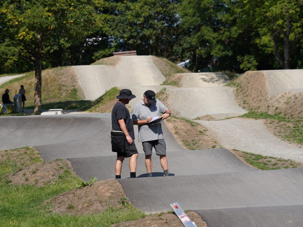 Zwei Männer stehen auf einem asphaltierten Pumptrack und notieren Daten auf einem Klemmbrett. Im Hintergrund sind die Hügel eines Bikeparks zu sehen.
