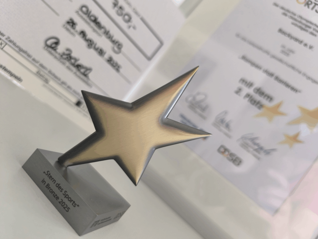 Ein bronzefarbener Stern-Pokal mit der Gravur „Stern des Sports in Bronze 2025“. Im Hintergrund verschwommen die Urkunden mit Unterschriften.