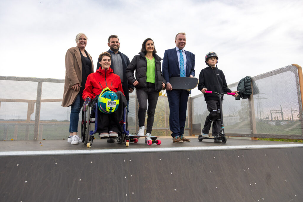 Mehrere Personen, darunter eine Person im Rollstuhl, stehen gemeinsam auf der neuen Miniramp in Bad Lippspringe und lächeln in die Kamera. Im Hintergrund ist der Himmel leicht bewölkt.