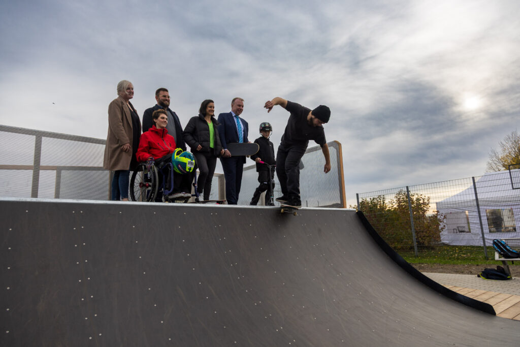 Gruppenfoto auf der Miniramp: Eine Person im Rollstuhl mit Helm in der Hand, daneben mehrere Erwachsene, ein Skateboarder und ein Kind mit Scooter. Ein Skateboarder fährt gerade die Rampe hinunter.