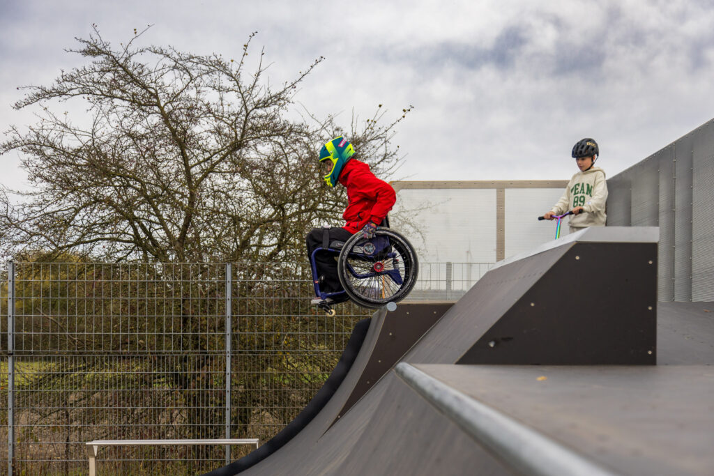 Eine Person im Rollstuhl mit roter Jacke und buntem Helm fährt die Miniramp in Bad Lippspringe hinunter. Rechts daneben steht ein Kind mit Scooter und Helm und schaut zu.