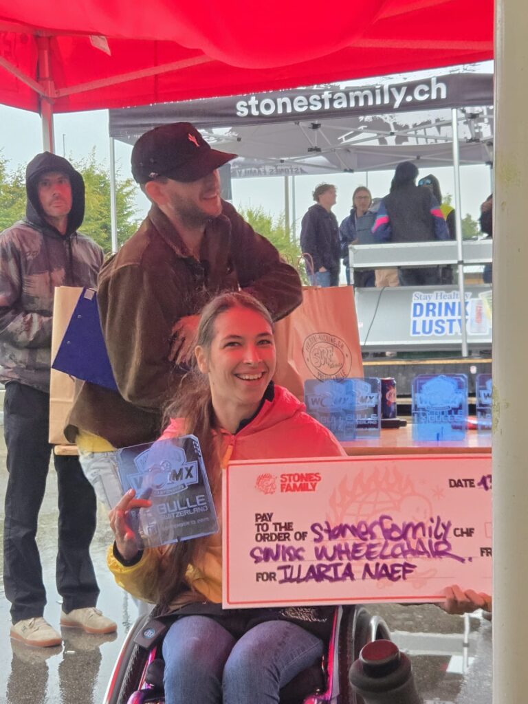 Eine Rollstuhlfahrerin hält im Regen unter einem Pavillon eine große Siegerscheck-Tafel in den Händen. Sie lächelt, während hinter ihr ein Moderator ins Mikrofon spricht.