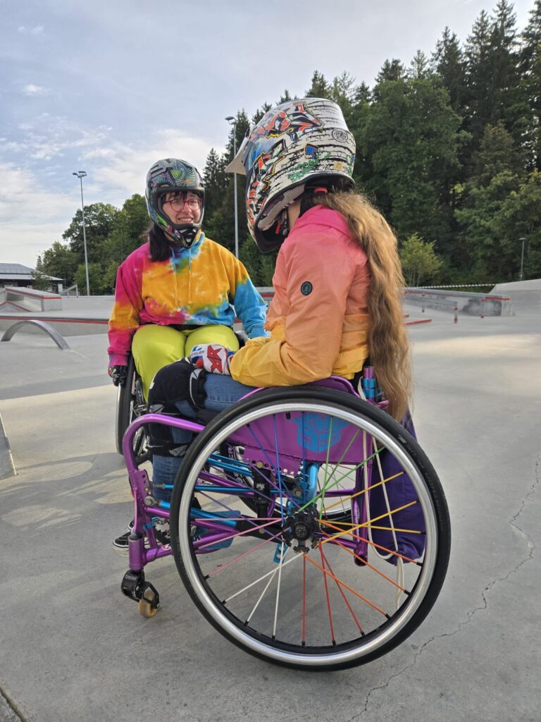 Zwei Rollstuhlfahrerinnen mit bunten Helmen und Regenjacken in leuchtenden Farben unterhalten sich lächelnd im Skatepark.