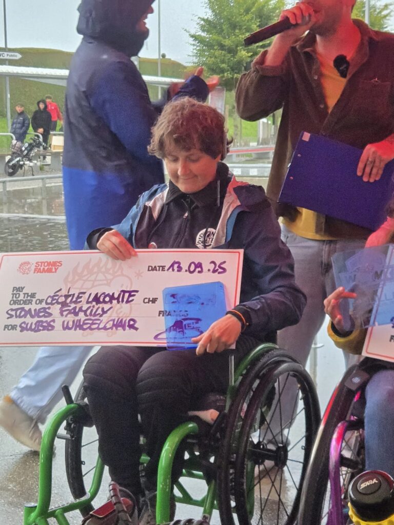 Eine Rollstuhlfahrerin mit langen Haaren hält im Rollstuhl sitzend ihre Trophäe und einen großen Siegerscheck.