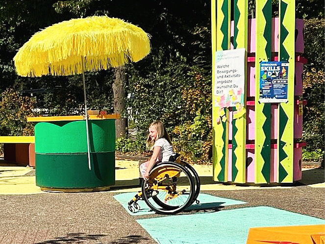Eine junge Person sitzt im gelben Rollstuhl und rollt über einen farbig markierten Weg. Um sie herum bunte Installationen mit einem großen gelben Sonnenschirm und bunten Holzsäulen. Im Hintergrund hängen Plakate mit Infos zur Beteiligungswerkstatt.