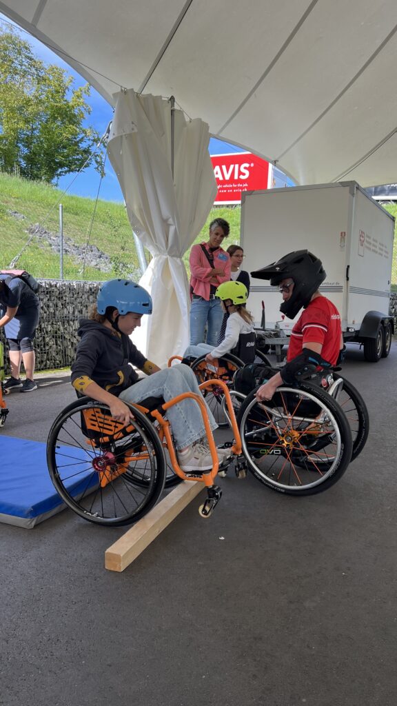 „Zwei Kinder im Rollstuhl mit Helm üben das Fahren über eine Holzrampe während eines WCMX-Workshops in Nottwil. Im Hintergrund stehen Begleitpersonen und das Team.“