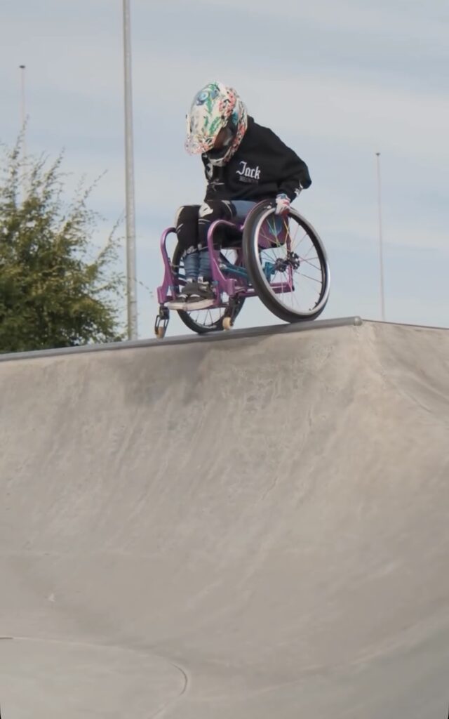 Eine Rollstuhlsportlerin mit buntem Helm steht oben auf einer hohen Rampe im Skatepark. Sie bereitet sich konzentriert auf den Drop-in vor.