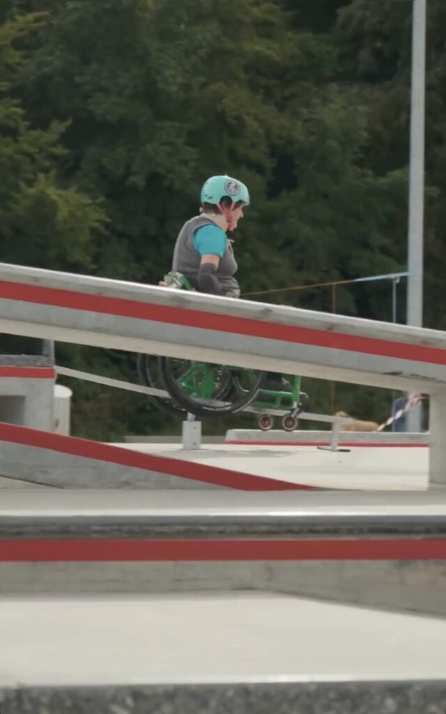 Eine Rollstuhlfahrerin mit türkisfarbenem Helm fährt über ein Rail im Skatepark. Hinter ihr sind Bäume und Zuschauer zu erkennen.