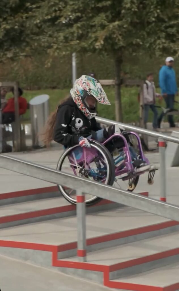 Eine Rollstuhlfahrerin mit buntem Helm und lila Rollstuhl fährt über eine Rampe mit roten Kanten.