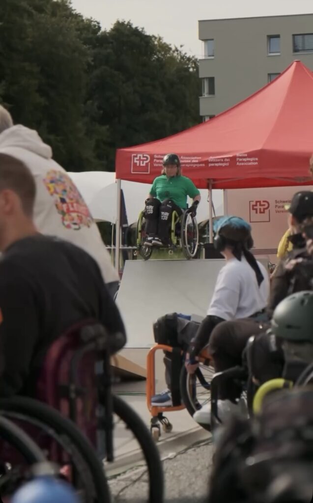 Eine Rollstuhlfahrerin mit grünem Shirt fährt die Rampe im Skatepark hinunter. Im Hintergrund sind Zuschauende und ein Pavillon zu sehen.