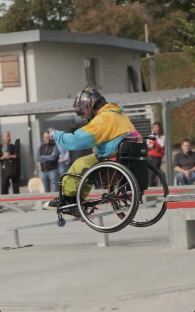 Eine Rollstuhlfahrerin mit bunter Jacke und Helm rollt über ein Rail im Skatepark, während Zuschauer im Hintergrund zusehen.