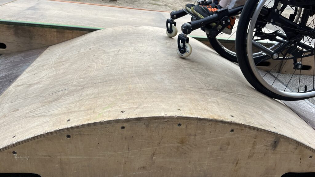 Nahaufnahme der Vorderräder eines Rollstuhls auf einer Holzrampe. Das Foto zeigt die Bewegung und den Kontakt der Räder mit der Oberfläche.
