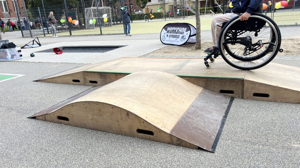 Ein Rollstuhl fährt über eine kleine Holzrampe. Im Hintergrund stehen ein Workshop-Banner und bunt geschmückte Bäume auf dem Gelände.