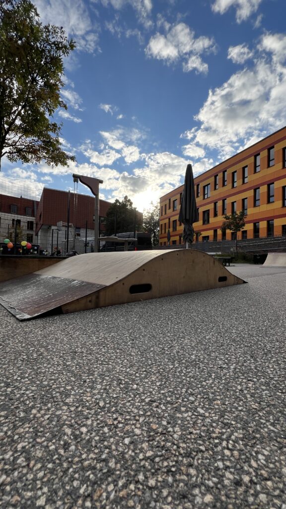 Eine hölzerne Skate-Rampe steht auf einem Innenhof. Die Sonne scheint durch die Wolken, dahinter sind bunte Gebäude des Oberlinhauses zu sehen.