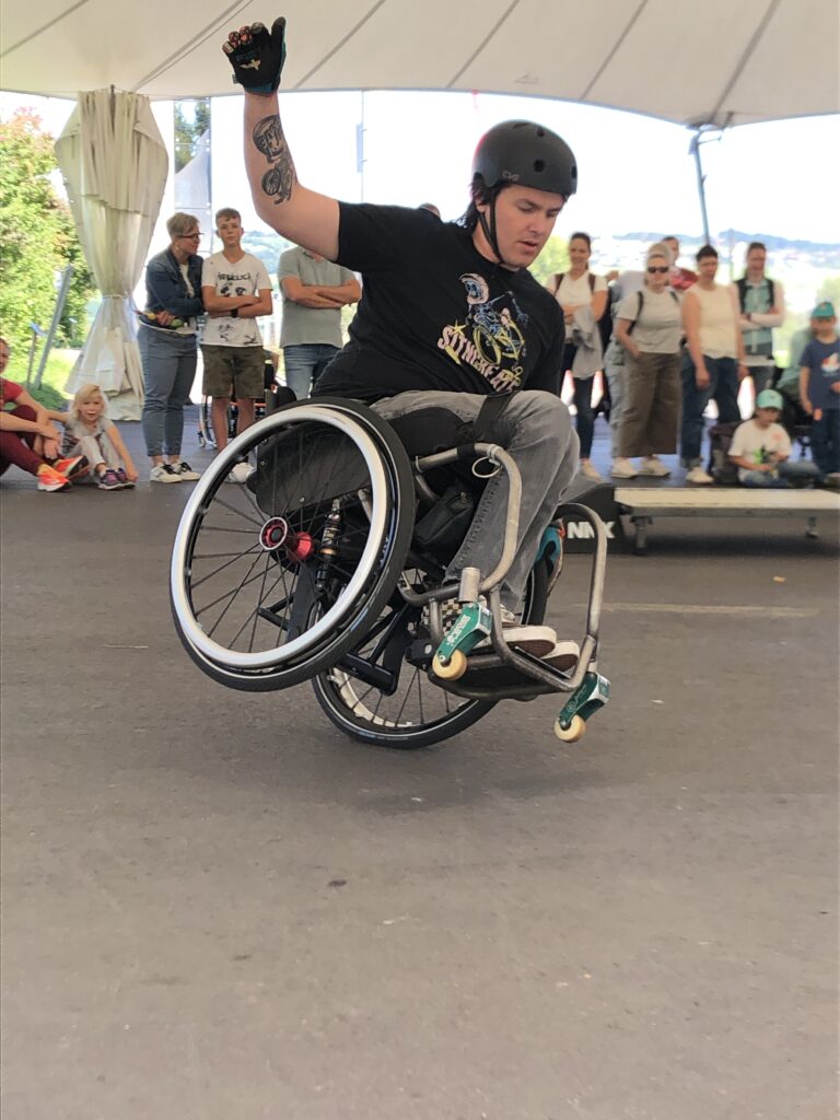„David Lebuser zeigt einen dynamischen Side-Wheelie mit seinem WCMX-Rollstuhl während der Show in Nottwil. Er trägt Helm und Handschuhe, das Publikum schaut gespannt zu.“