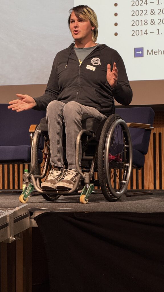 „David Lebuser sitzt im Rollstuhl auf einer Bühne, spricht gestikulierend während seiner Präsentation beim inklusiven Sportkongress ‚Sport verbindet‘ im Campus Sursee. Im Hintergrund ist eine Präsentationsfolie mit Stichpunkten zu sehen.“