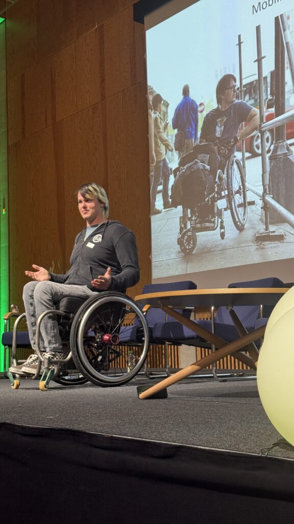 „David Lebuser hält seinen Vortrag auf der Bühne in Sursee. Im Hintergrund ist ein großes Bild projiziert, das ihn früher im Alltag zeigt – als Kontrast zum Thema Mobilität und Teilhabe.“