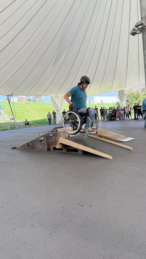 „David Lebuser fährt mit seinem WCMX-Rollstuhl eine Rampe bei einem Outdoor-Workshop im Schweizer Paraplegikerzentrum in Nottwil herunter. Er trägt einen Helm, Zuschauende stehen im Hintergrund unter einem weißen Zeltdach.“