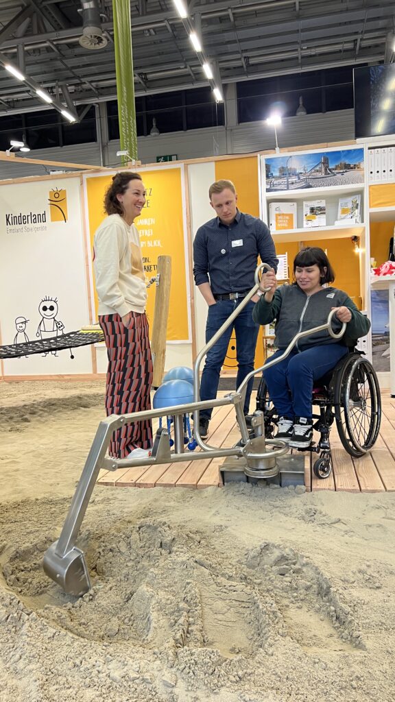 Lisa, eine Frau im Rollstuhl bedient auf einer Messe einen silbernen Sandbagger mit zwei Griffen. Zwei Personen stehen daneben und beobachten lächelnd. Der Bagger gräbt Sand aus einer kleinen Sandfläche, die auf Podestholz eingefasst ist. Im Hintergrund ein Messestand mit gelben Wänden und der Aufschrift „Kinderland“.
