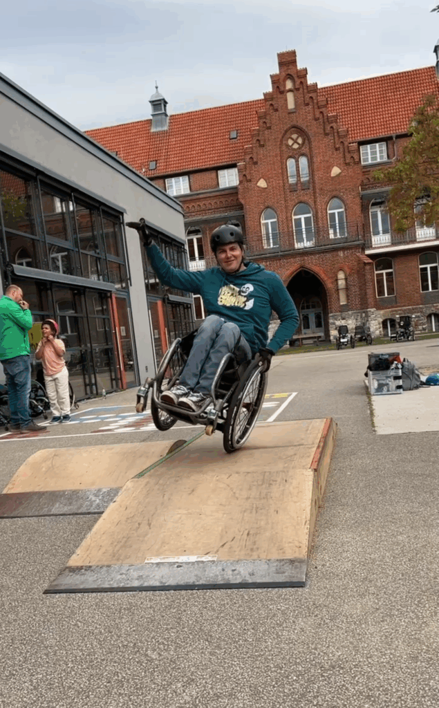 Eine Person im Rollstuhl trägt Helm und Hoodie und rollt mit Schwung eine kleine Rampe hinunter. Im Hintergrund sind ein rotes Backsteingebäude und weitere Personen zu sehen.