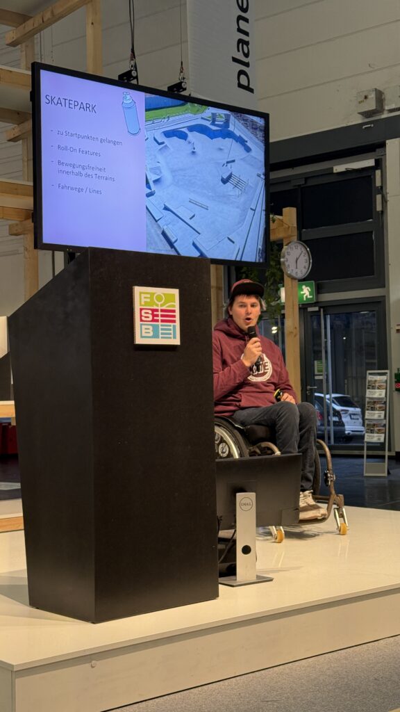 David sitzt im Rollstuhl auf einer Bühne und hält ein Mikrofon, während hinter ihm auf einem großen Bildschirm eine Skatepark-Grafik und Stichpunkte zu Rollstuhlzugängen angezeigt werden. Vor ihm steht ein großes Rednerpult mit dem FSB-Logo.