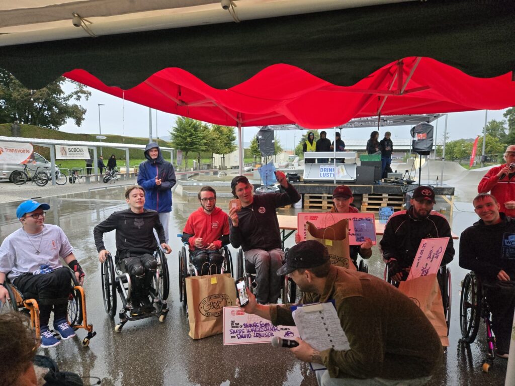 Siegerehrung der Rollstuhlfahrenden Männer unter einem provisorisch aufgebautem Pavillon . Im Hintergrund ein verregneter nasser Skatepark. 