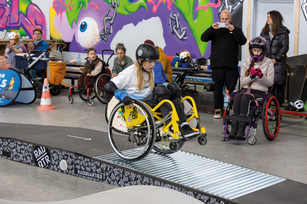 Ein Kind im gelben WCMX-Rollstuhl fährt konzentriert über eine schmale Rampe mit Rollen, die von Radquartier gesponsert wurde. Andere Kinder und Erwachsene im Rollstuhl sowie Zuschauende sitzen im Hintergrund vor einer bunten Graffiti-Wand und beobachten den Run.