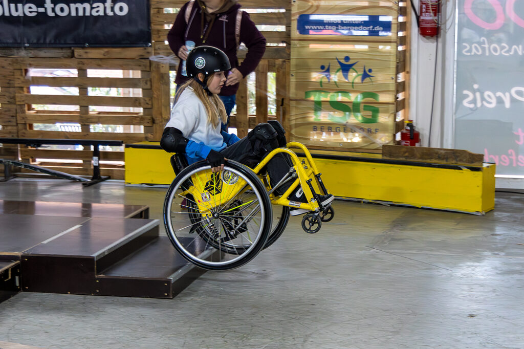 Ein Kind im gelben WCMX-Rollstuhl fährt eine kleine Treppe in der Skatehalle Allerfornia hinunter. Das Kind trägt Helm und Schutzausrüstung und hält den Rollstuhl leicht im Wheelie. Im Hintergrund stehen Personen und Elemente der Skatehalle, darunter ein Banner und ein Schild der TSG Bergedorf.