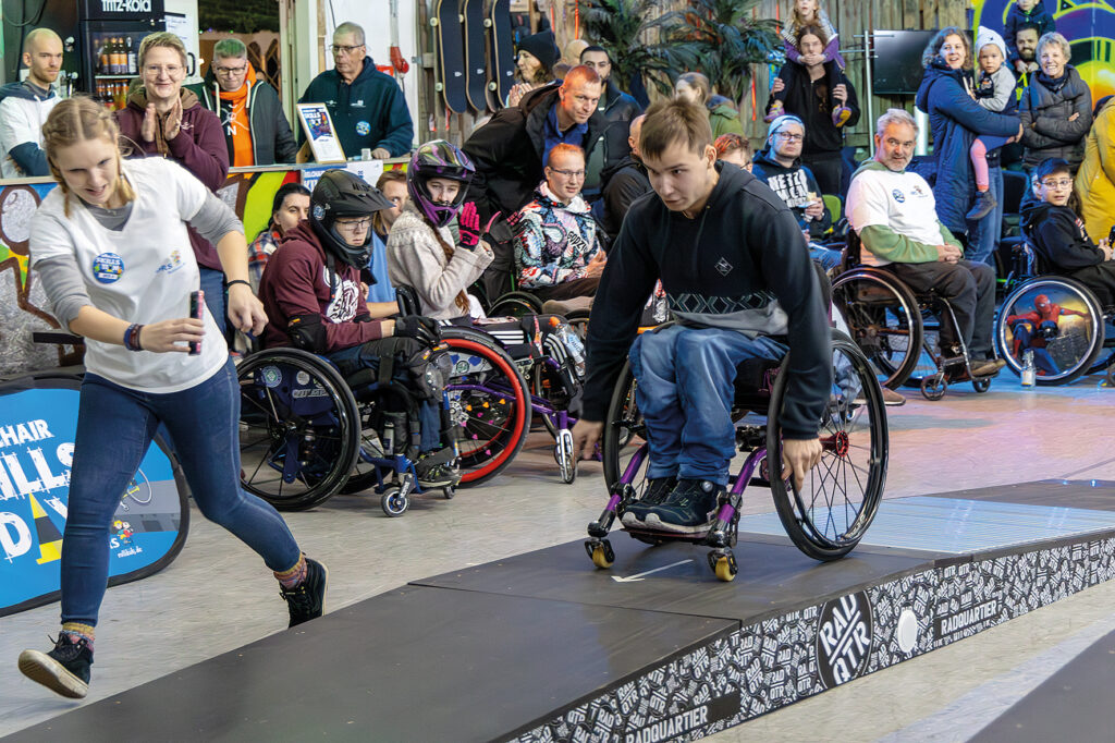 Ein Jugendlicher fährt im WCMX-Rollstuhl dynamisch eine Rampe von Radquartier hinunter, während eine Helferin daneben mitläuft und die Zeit misst. Im Hintergrund sitzen und stehen viele Zuschauende und Teilnehmende in der Skatehalle.