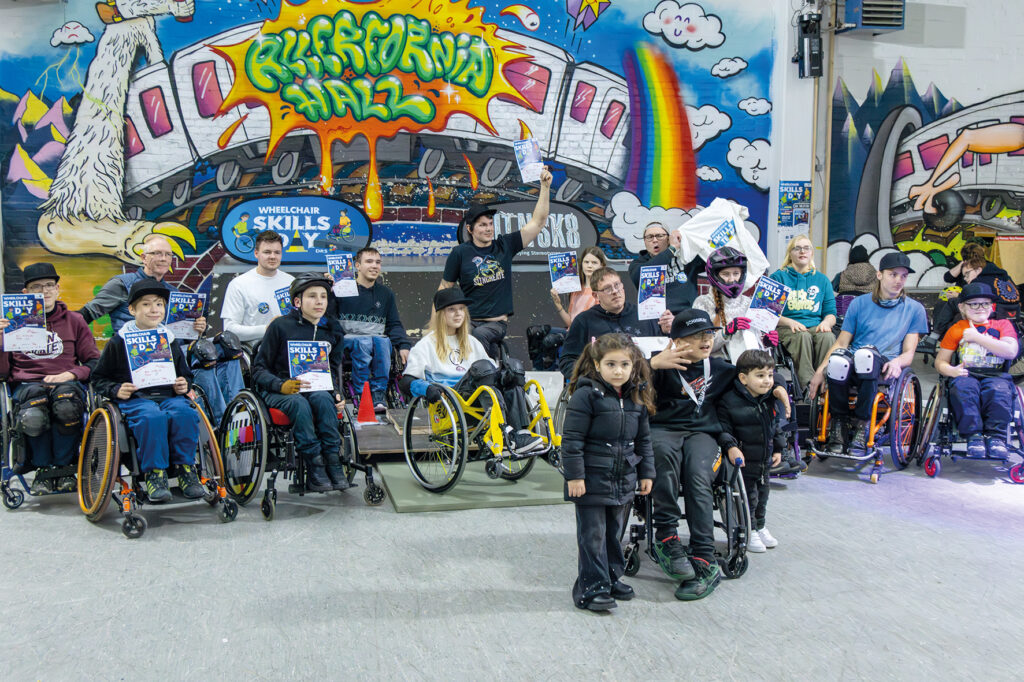 Gruppenfoto aller Teilnehmenden des Wheelchair Skills Day. Menschen im Rollstuhl und einige Kinder stehen oder sitzen vor einer bunt bemalten Graffiti-Wand in der Allerfornia Skatehalle. Viele halten ihre Urkunden hoch und lächeln in die Kamera.