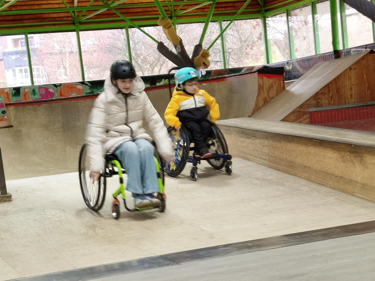 Zwei Menschen im Rollstuhl fahren nebeneinander eine flache Rampe herunter. Links eine erwachsene Person mit hellem Mantel und Helm, rechts ein Kind mit blauem Helm und gelber Jacke. Beide bewegen sich dynamisch Richtung Kamera. Der Skatepark ist hell und weitläufig.