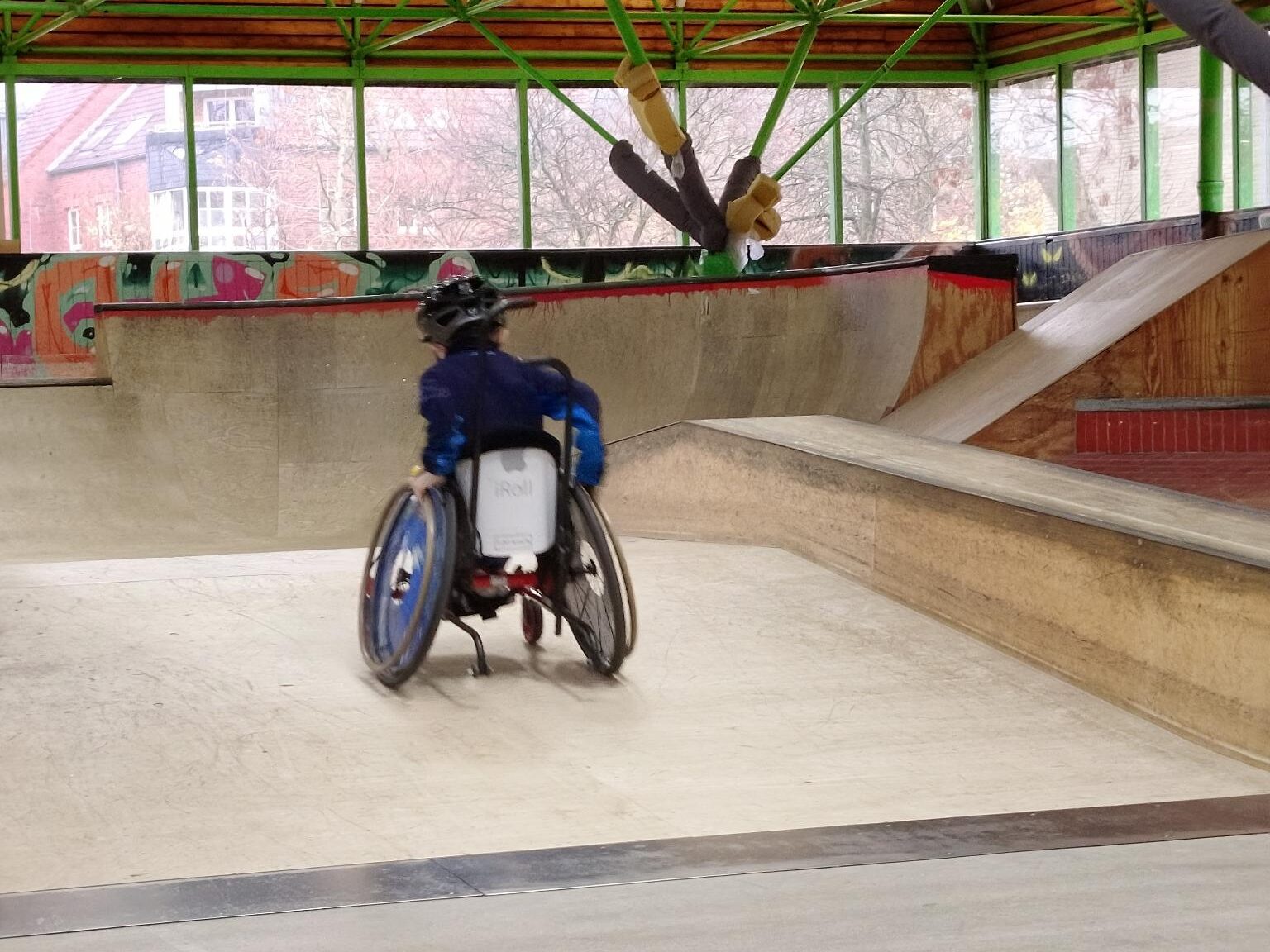 Eine Person im Rollstuhl mit blauem Helm und dunkler Kleidung fährt eine Rampe im Skatepark rauf. Der Rollstuhl hat eine weiße Rückenplatte mit der Aufschrift „iRoll“. Im Hintergrund sind weitere Rampen, Graffiti und große Fenster mit Blick ins Freie.