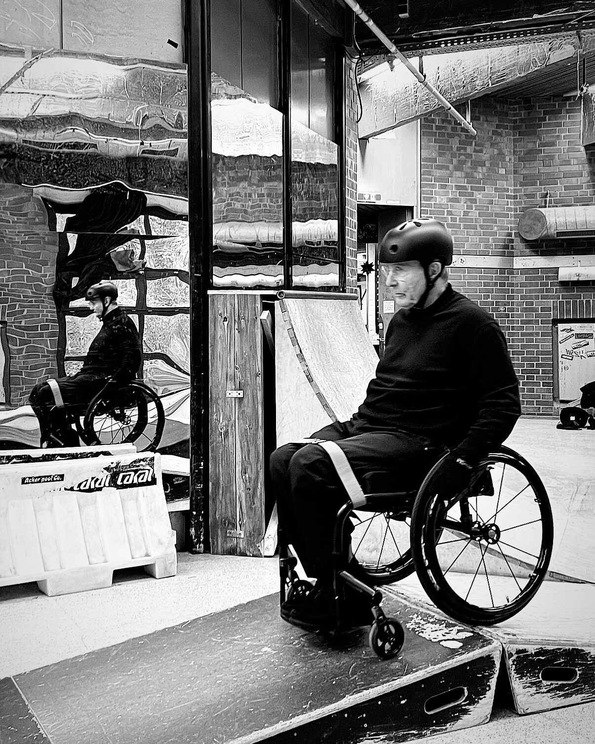Schwarz-weiß-Foto eines älteren Mannes im Rollstuhl in einer Skatehalle. Er trägt einen Helm und sitzt auf einer kleinen Rampe, den Blick leicht nach unten gerichtet. Hinter ihm spiegeln sich Rampen und sein eigenes Bild in einer verzerrten Spiegelfläche. Im Hintergrund stehen eine Quarterpipe und eine Backsteinwand. Die Szene wirkt ruhig und konzentriert.