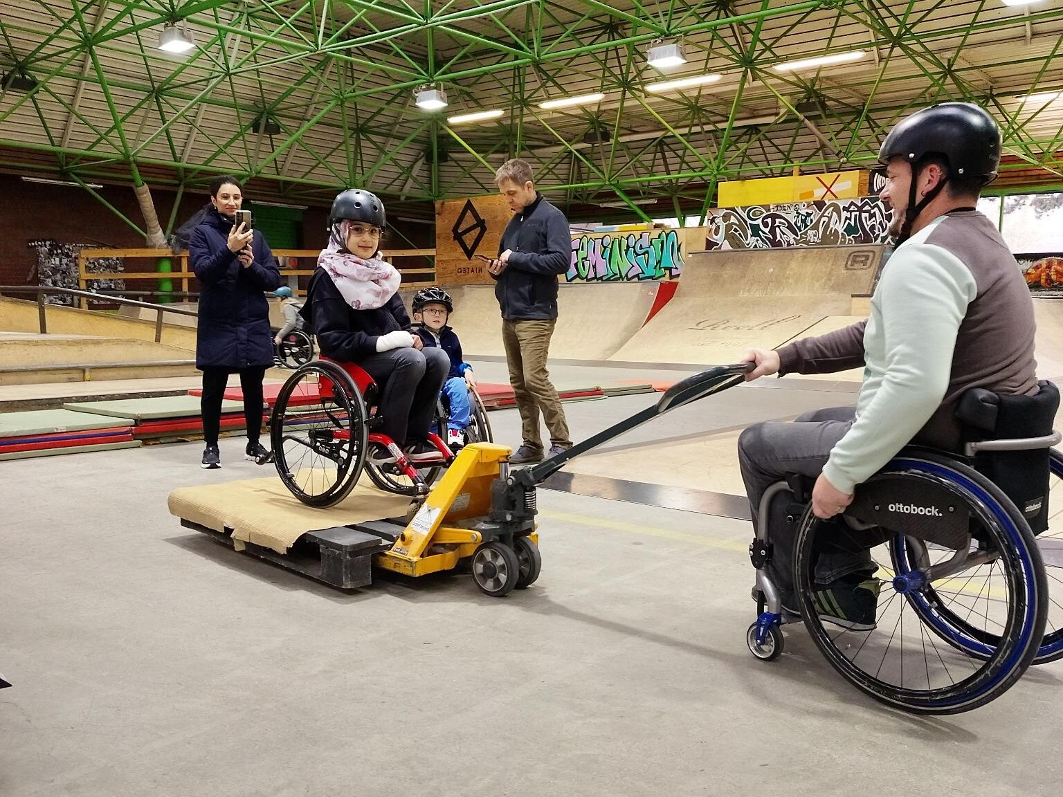In einer Skatehalle steht eine junge Person im Rollstuhl auf einer Holzplattform, die mit einem Hubwagen bewegt wird. Eine weitere Person im Rollstuhl zieht den Hubwagen. Im Hintergrund beobachten zwei Erwachsene die Szene, einer davon hält ein Smartphone. Überall sind Rampen, Graffiti und grüne Dachstreben sichtbar.
