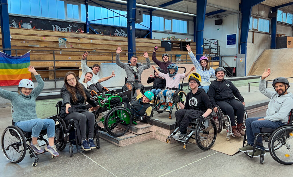 Ein Gruppenfoto in einer Skatehalle mit vielen Rollstuhlfahrenden Menschen von jung bis alt. Sie jubeln, einige liegen zurückgelehnt auf Stufen. Im Hintergrund sind Skaterampen zu sehen und eine Regenbogenfahne.