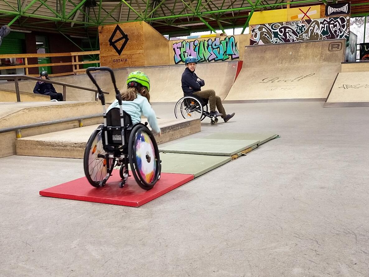 Mehrere Personen im Rollstuhl bewegen sich durch eine große Skatehalle. Im Vordergrund fährt ein Kind mit grünem Helm über eine rote Matte, im Hintergrund rollt ein Erwachsener eine Rampe hinunter. Die Halle ist weitläufig, mit vielen Holzrampen, grünen Stahlträgern und Graffiti an den Wänden.