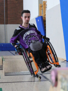 Ein Rollstuhlfahrer auf die Seite gelehnt fährt durch den mobilen Skatepark in einer Sporthalle. 