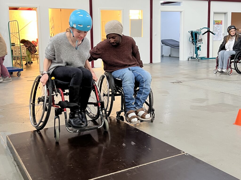 Eine weiße Frau im Rollstuhl versucht eine Kante an unserem Rollstuhlparcours zu überwinden. Ein Schwarzer Rollstuhlfahrender Übungsleiter hilft dabei.