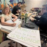 Mehrere junge Menschen sitzen in einem kreativ gestalteten Innenraum an einem Tisch und malen Schilder für eine Protestaktion. Zwei der Personen sitzen im Rollstuhl. Im Vordergrund liegt ein großes selbstgemaltes Schild mit der Aufschrift: „Mobilität = nice to have = Grundrecht“. Auf dem Tisch stehen Pinsel, Stifte, Gläser und Farben.