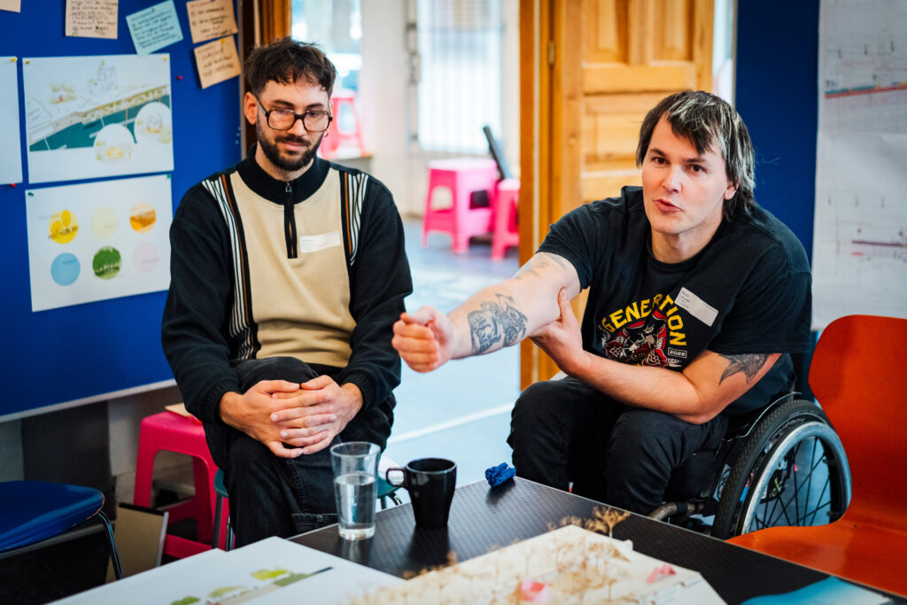 David Lebuser sitzt in seinem Rollstuhl an einem Tisch und spricht mit ausgestrecktem Arm in eine Diskussionsrunde. Neben ihm sitzt ein weiterer Teilnehmer. Im Hintergrund hängen Skizzen und Notizen zur Planung der Hafenkante.