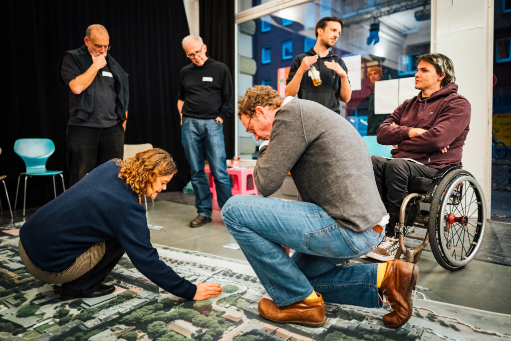 Mehrere Personen beugen sich über einen großen Luftbild-Plan der Hafenkante und diskutieren einzelne Bereiche. Rechts sitzt David Lebuser im Rollstuhl und beteiligt sich am Gespräch. Die Szene zeigt die gemeinsame Arbeit an konkreten Planungsideen.